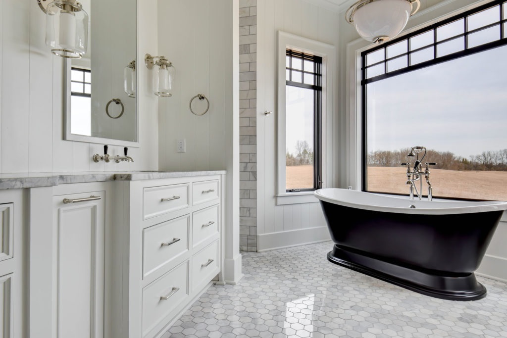 Interior of a spacious bathroom with a classic design, featuring a freestanding black bathtub with silver fixtures near a large window overlooking a field. The room has white walls and cabinetry with a marble countertop and dual sinks. Two glass pendant lights hang above the vanity, and the floor is tiled with white hexagonal tiles.